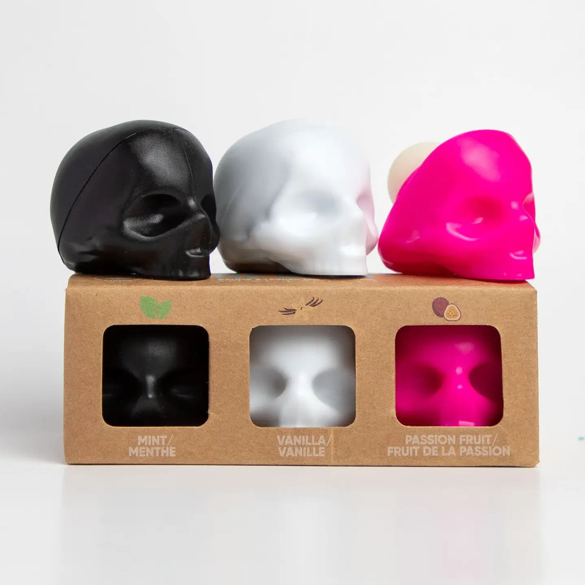 Three skull-shaped lip balms in matte black, pearlescent white, and hot pink displayed on and in front of a kraft cardboard gift box labeled mint, vanilla, and passion fruit.