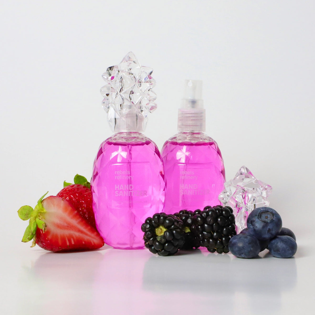 Two hot pink faceted pineapple-shaped hand sanitizer spray bottles by Rebels Refinery, one with a decorative clear star-cluster cap and one with a spray nozzle, surrounded by fresh strawberries, blackberries, and blueberries on a white surface.