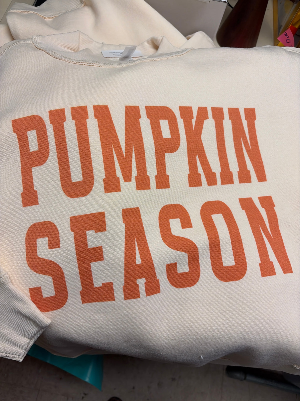 A folded ivory pullover sweatshirt with a bold orange varsity-font graphic reading "Pumpkin Season" across the chest and a ribbed mockneck collar.