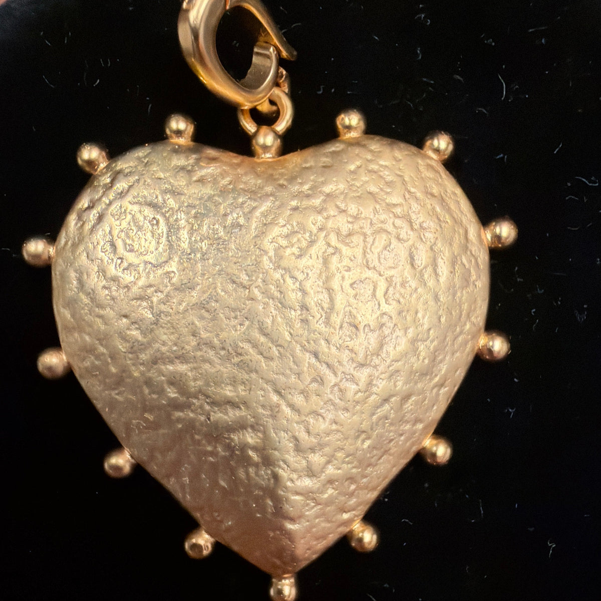 A textured gold heart-shaped pendant with small gold studs around the edge and a gold lobster clasp at the top, displayed on a black circular velvet surface.