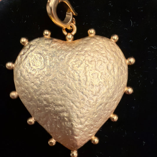 A textured gold heart-shaped pendant with small gold studs around the edge and a gold lobster clasp at the top, displayed on a black circular velvet surface.