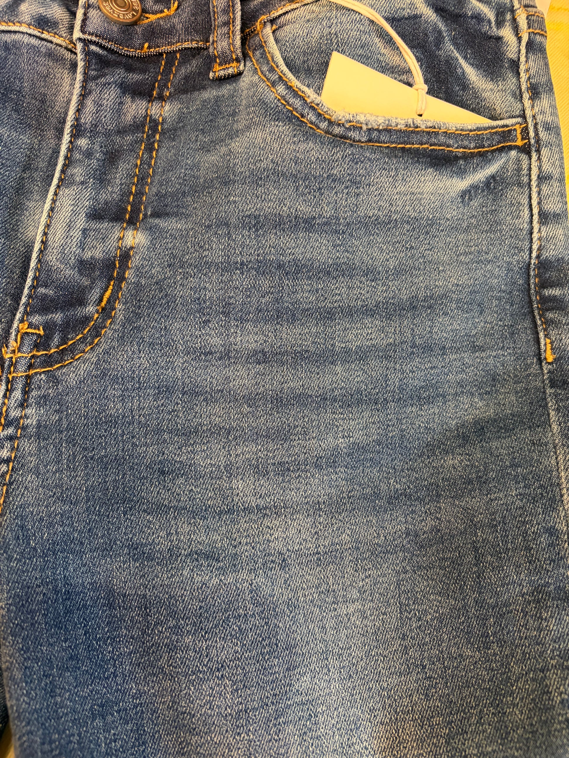 Vervet Masterfully Jean jeans hanging on a wooden background, featuring classic denim style and detailed stitching.