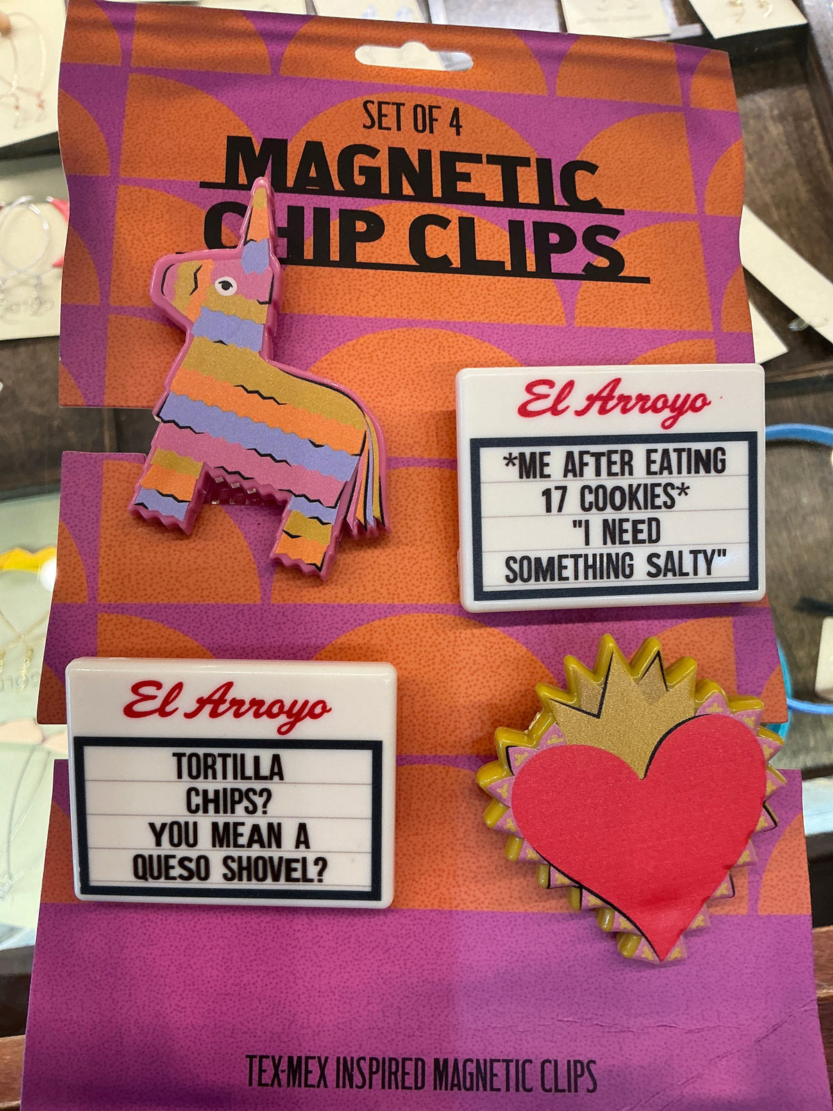 A set of four El Arroyo magnetic chip clips displayed on their original packaging. The clips include a colorful pinata, a red heart with gold jagged edges, a taco, and a white rectangular clip with humorous text prompts, all resting on a purple and orange patterned card.